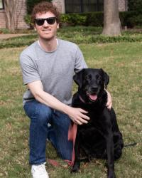 Image of a white man with sunglasses on kneeled beside his black lab dog