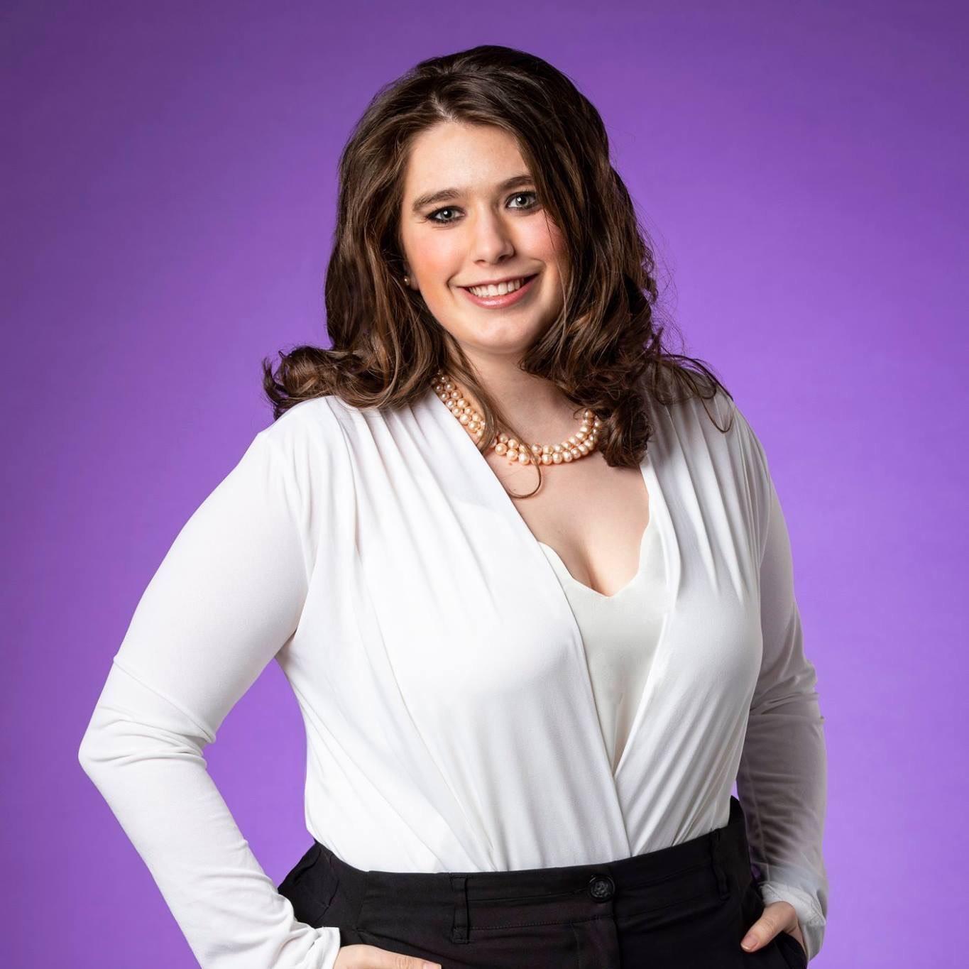 Ashley smiling, hands in pockets, wearing a white top and necklace, standing in front of a purple background.