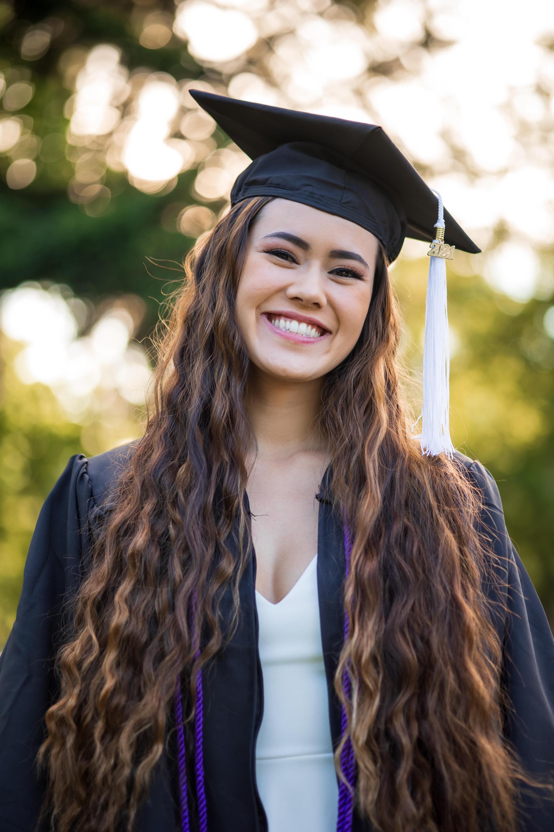Closeup image of an Asian American woman with long dark brown wavy hair, wearing a white dress, Black graduation gown and cap with a white tassel, and purple cords hanging from her neck, smiling. Background is blurred greenery.