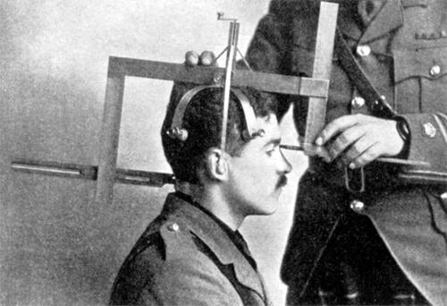 white man seated with a measuring device on his head, held by a white man wearing a uniform