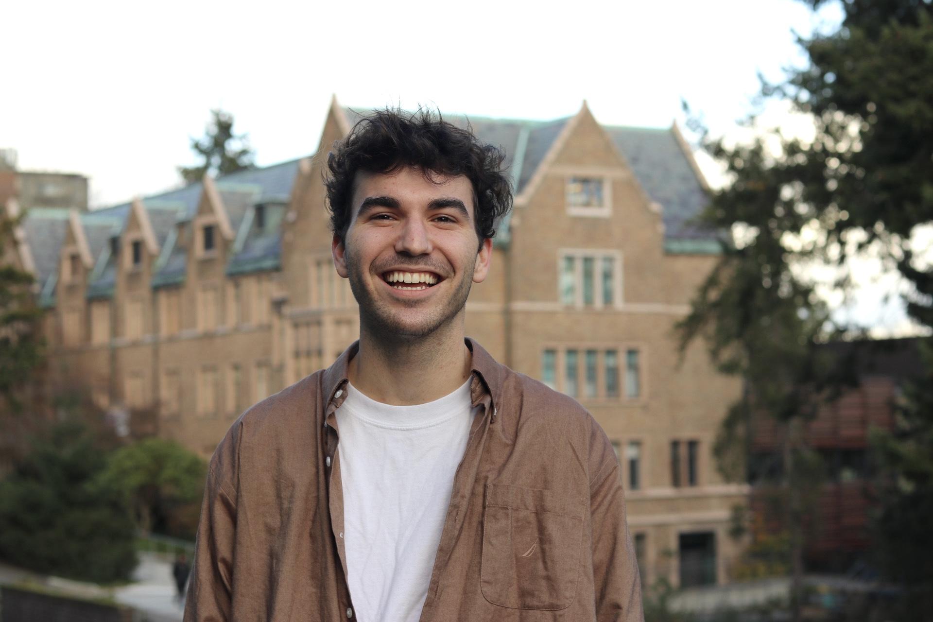 Closeup image of a white man with short fluffy brown hair wearing a white t-shirt and brown collared shirt overtop, smiling in front of a UW building.