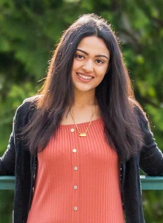 Ishira, a girl with straight black hair and brown eyes, is wearing an orangish pink dress with a black sweater and smiles at the camera.