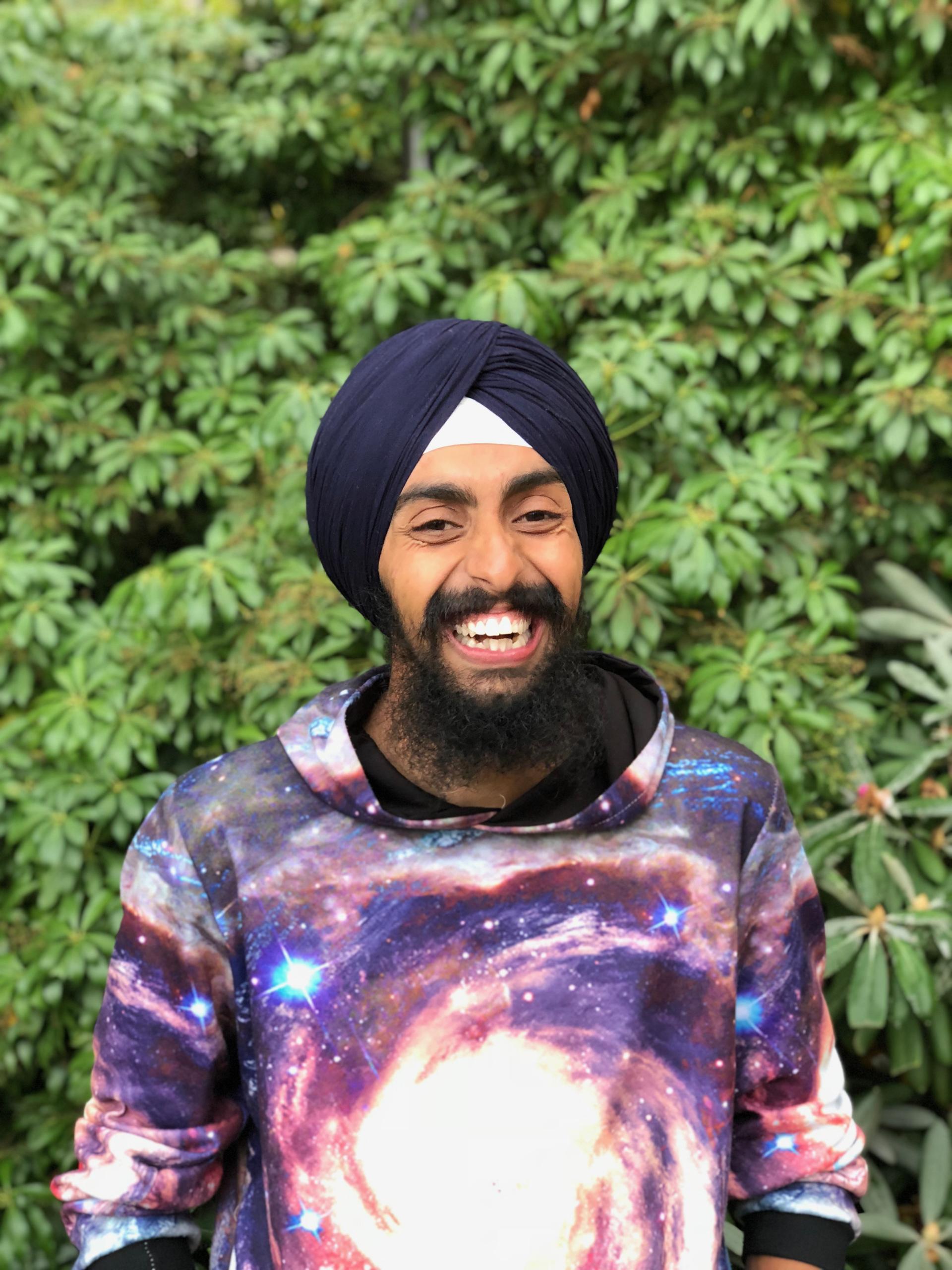 Jasmair smiling broadly, wearing a turban and brightly-colored shirt, standing in front of green foliage.