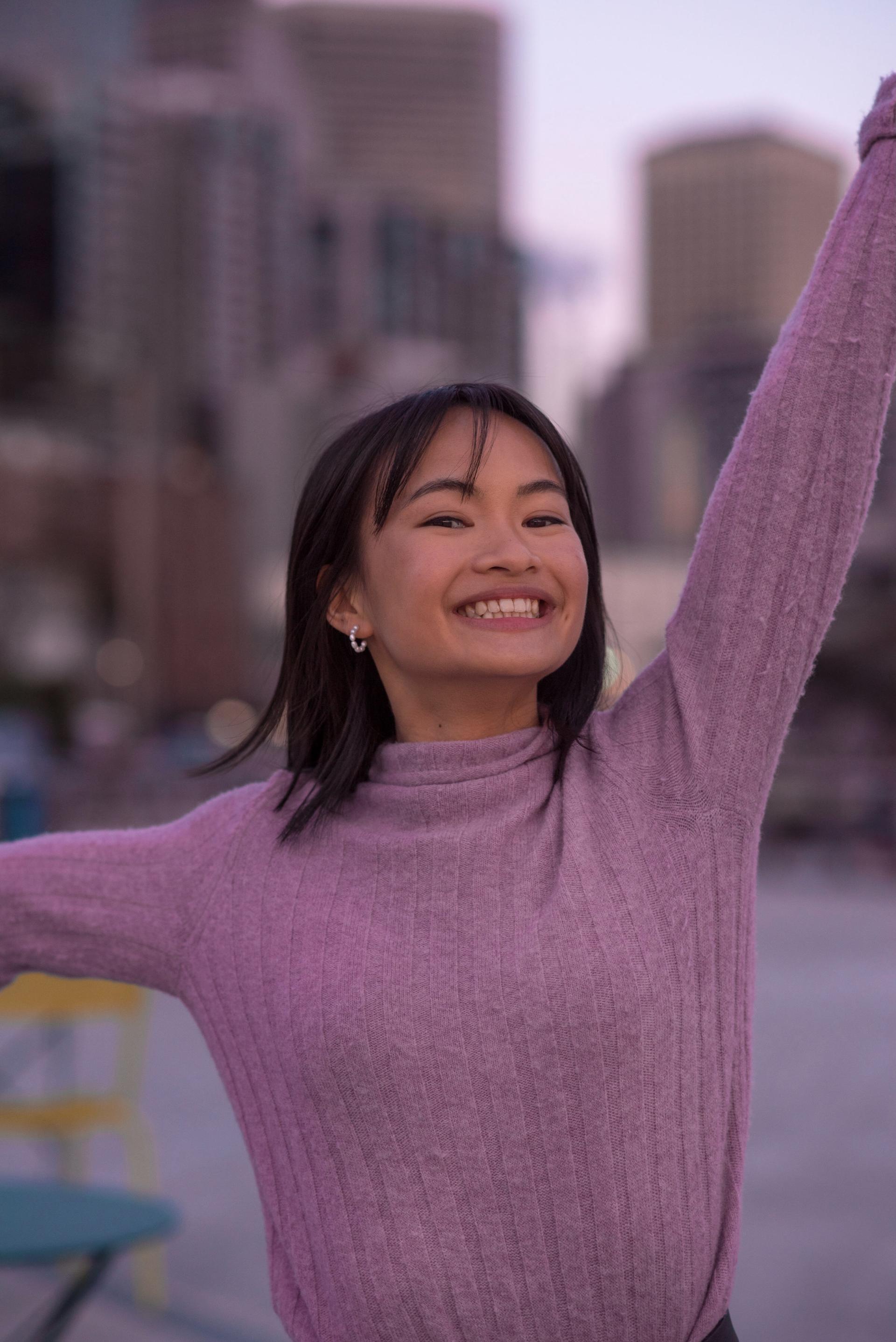 Katherine, Asian woman, short brown hair with bangs, wearing a pink sweater, smiles at the camera with her left arm raised up and her right arm to the side in front of out-of-focus buildings.