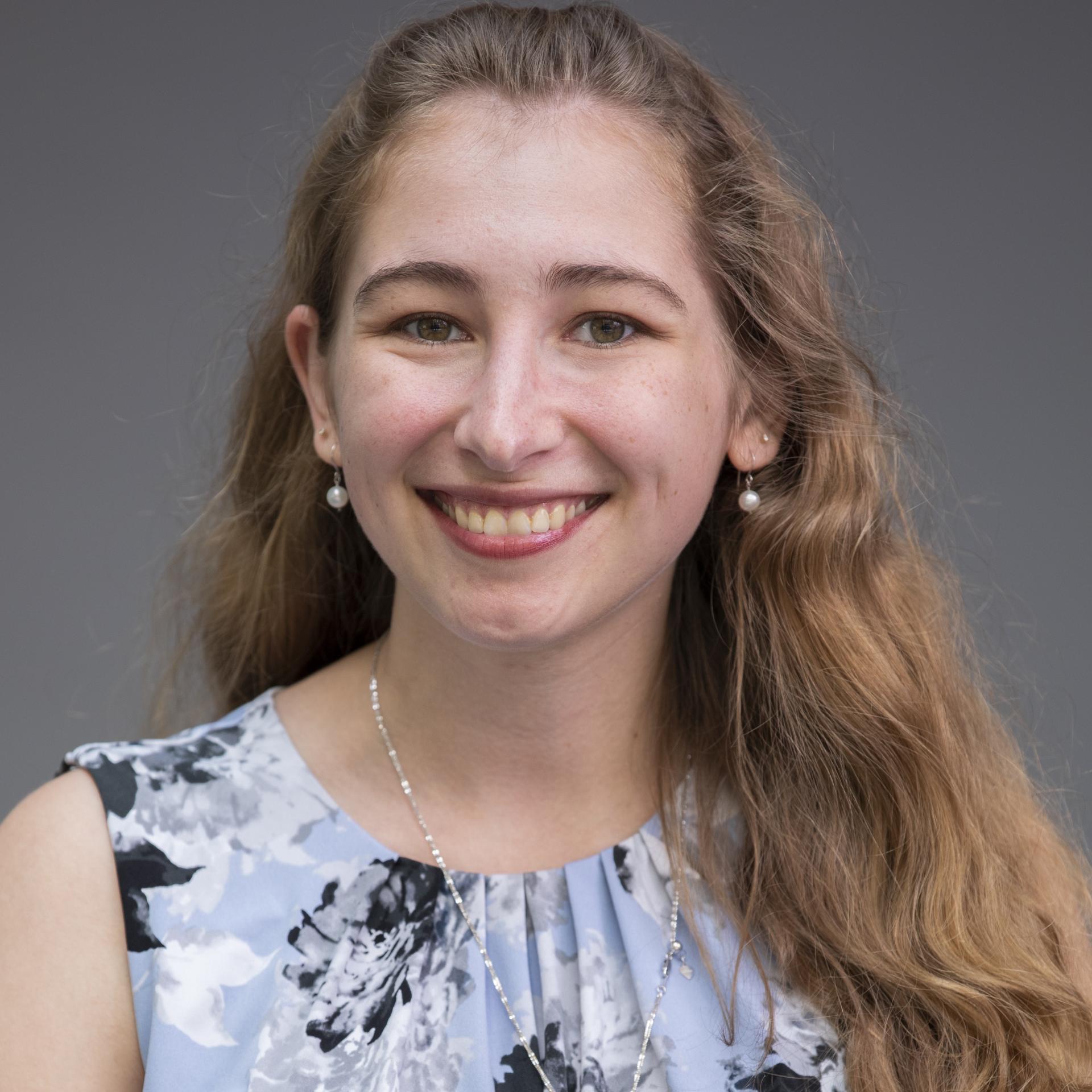 Kelly Mack, A young white woman with wavy brownish hair smiles for the camera. She wears a blue floral blouse.